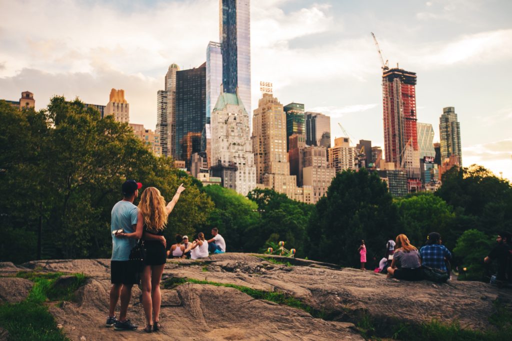 A view of Manhattan's downtown (all the skyscrapers, grey cloudy sky) from the rock formation in Central Park. A young couple with their backs to the camera point at the skyscrapers.