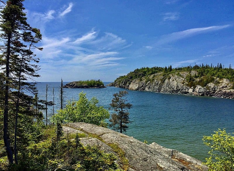 A bright blue cove surrounded by sloping grey hills and a tall pine tree, with shorter deciduous trees below. (Pukaskwa NP, Ontario)