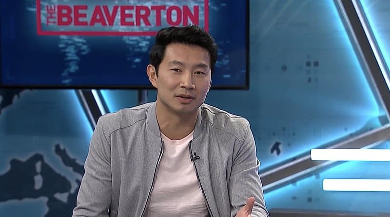 A young man on a mock-news set looks at the camera.