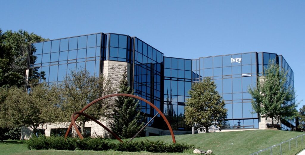 A multistorey glass building rests on a green hill, surrounded by trees and a wheel sculpture.