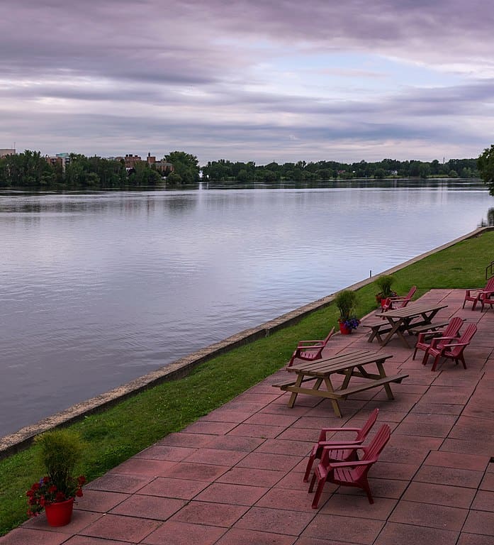 A quiet river flows through Laval, Quebec. A lawn and red brick patio with picnic tables is visible in the right side of the image.
