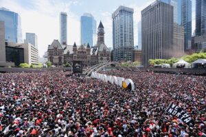 Toronto's downtown on We The North Day after the Raptors' win in 2019: tall steel and glass towers frame a sea of people standing in the public square in front of City Hall. Most are in red and black clothing.