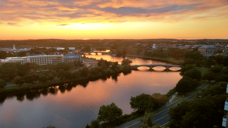 A view of the Harvard University campus at sunset: golden-red sky, winding river with a classic bridge, lots of trees and brick buildings.