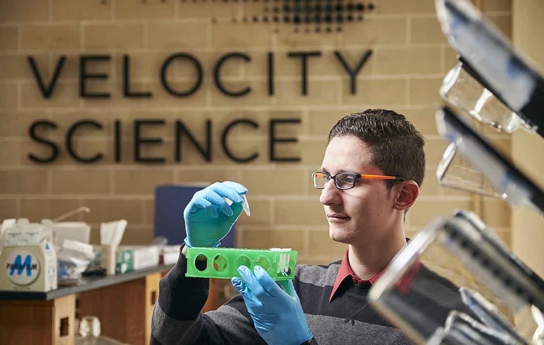 Student studying STEM, shown in a science classroom wearing latex gloves and looking at an eyedropper of liquid