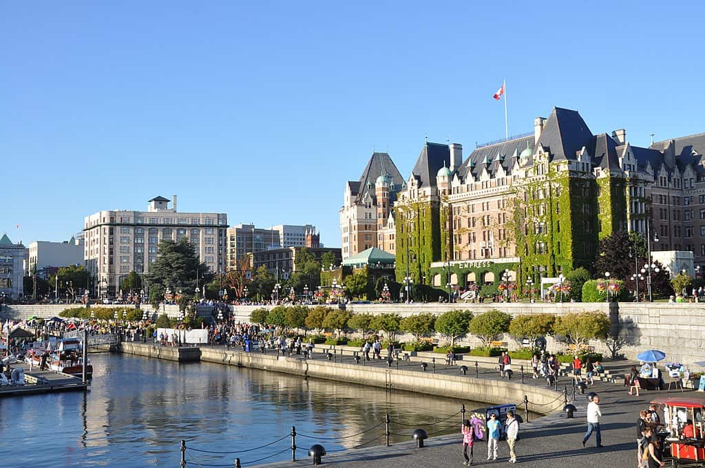 A tall brick hotel sits beside an urban waterfront with walking paths in Victoria, BC