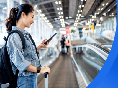 A female international student (in a Canadian tuxedo: a denim jacket and jeans!) smiles as she looks at her phone while standing on a moving walkway in an airport.