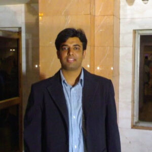 A smiling South Asian man in a blue suit stands next to a windown in an office. (RP Ankit Jain)