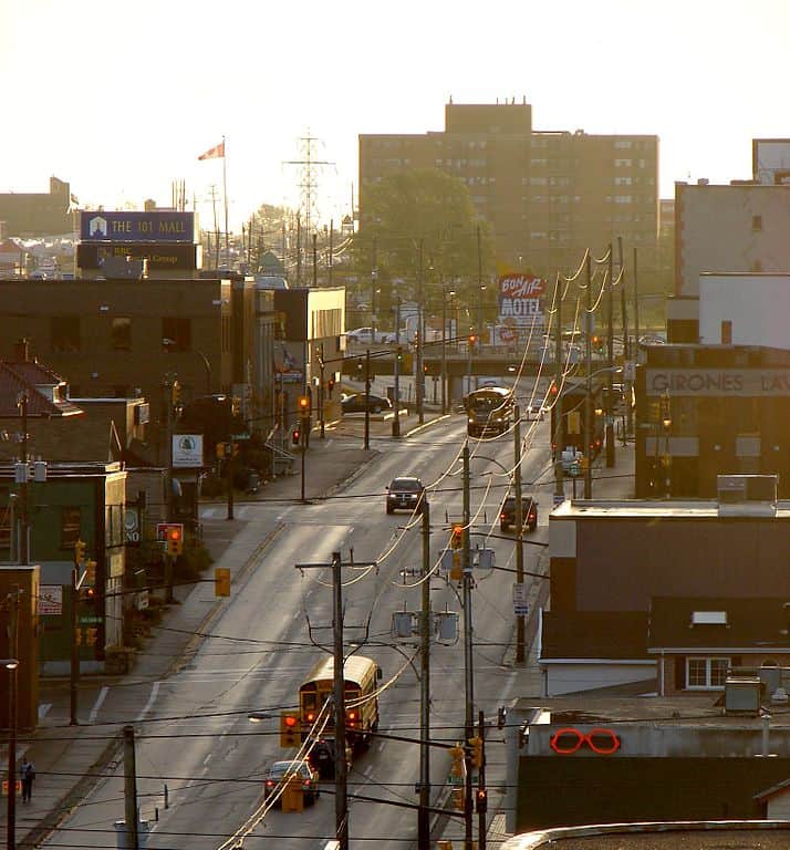 A four-lane road is flanked by medium-height brick buildings and street lights at dusk in Timmins, Ontario