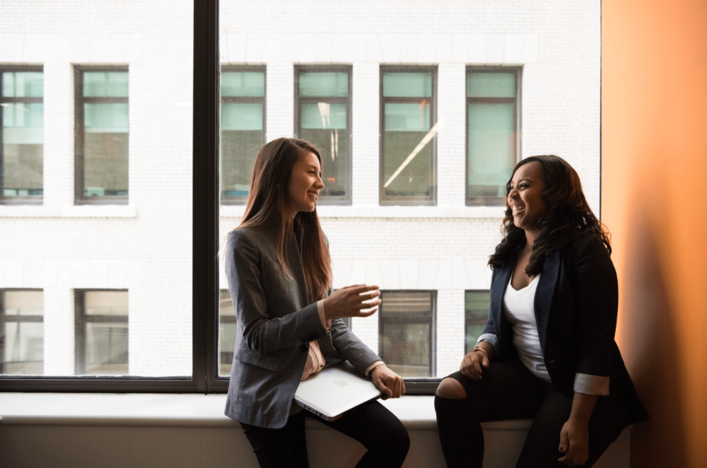 Two young women in business casual attire sit at a windowsill in an urban setting.