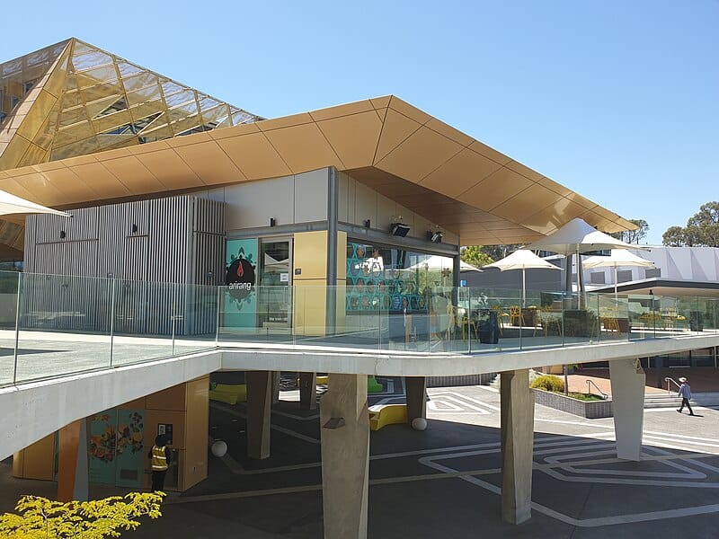 A modern multistorey concrete and glass building with large windows and a light tan roof on the Edith Cowan University Joondalup campus. The building appears on concrete "stilts" with a walkway extending to the lower left of the frame.