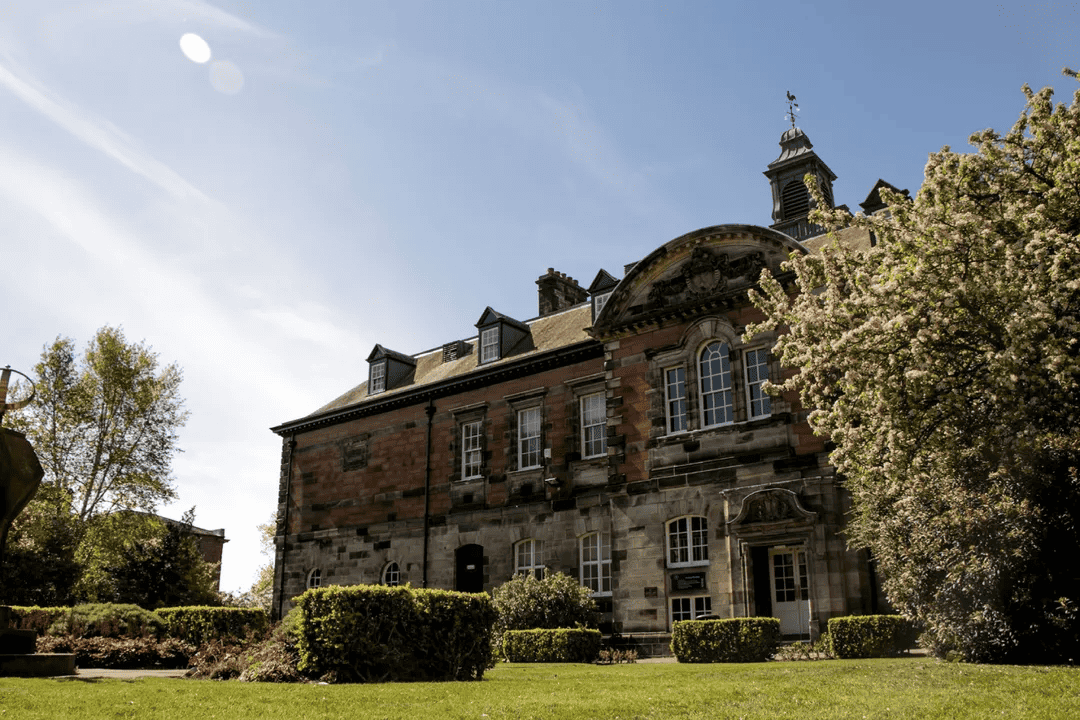 A rustic University of Dundee building before a blue sky with shrubbery in the foreground.