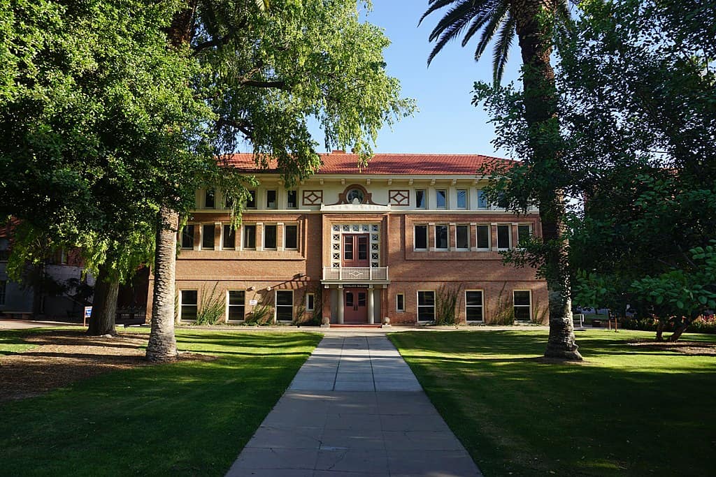 A Spanish colonial-style university building (red tile roof, warm brick) is framed by trees on the University of Arizona campus.