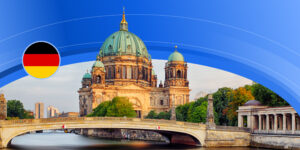 A boat sails under a bridge, which crosses a body of water somewhere in Germany. An impressive sand-coloured building with green copper domes sits behind the bridge.