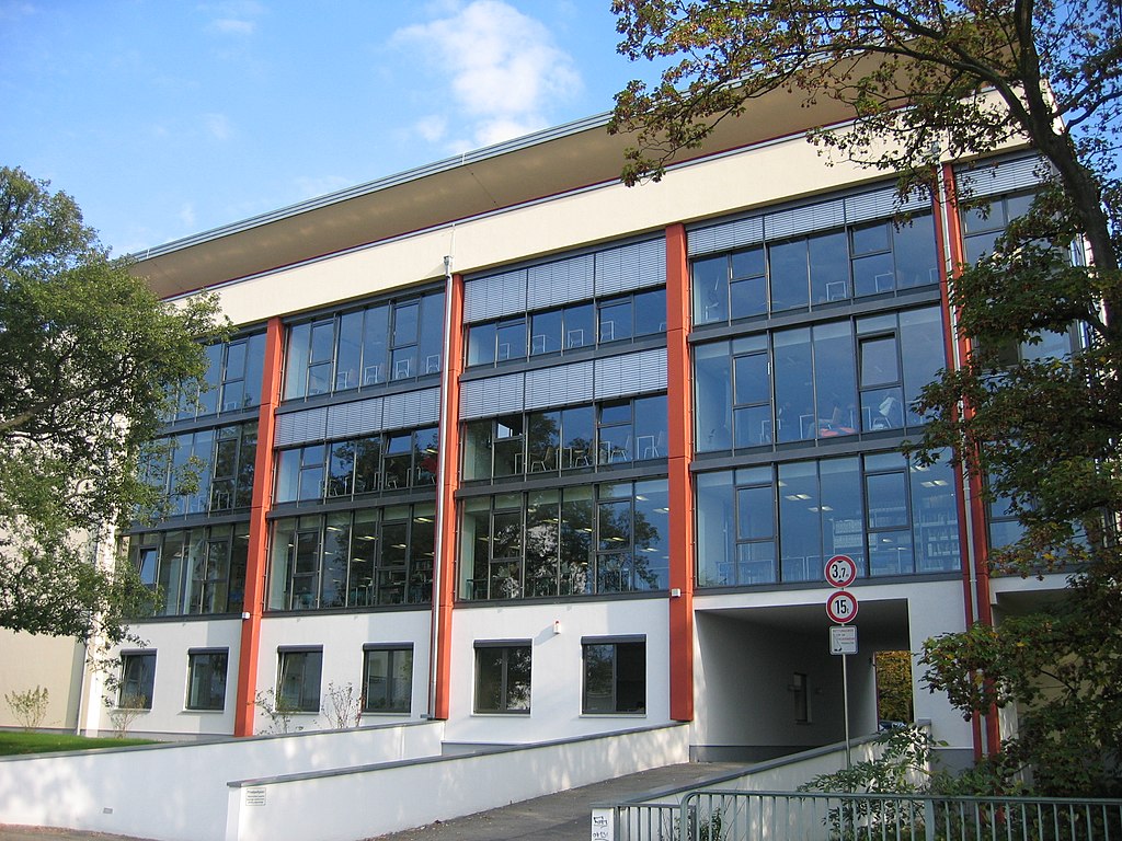 A midcentury academic building with glass sides on the IU (Bad Honnef) campus. The building is framed by a walkway and deciduous trees.