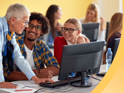 An instructor speaks with two international students in a sunny classroom, surrounded by other students.