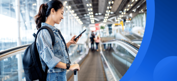 A female international student (in a Canadian tuxedo: a denim jacket and jeans!) smiles as she looks at her phone while standing on a moving walkway in an airport.