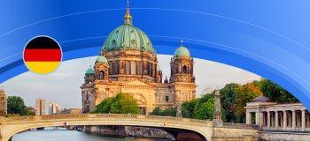 A boat sails under a bridge, which crosses a body of water somewhere in Germany. An impressive sand-coloured building with green copper domes sits behind the bridge.