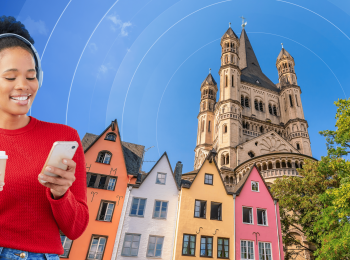 A smiling young woman looks down at her phone while holding a white to-go coffee mug. She is framed by a photographic background with colourful multistorey houses, a warm-hued stone castle, and a German flag logo.