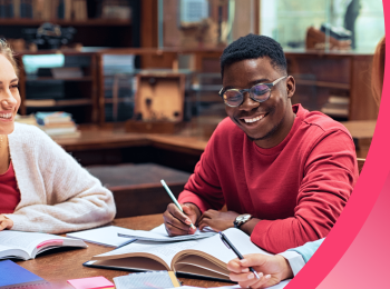 Two students sit at a table in a library, studying together and smiling with their books open